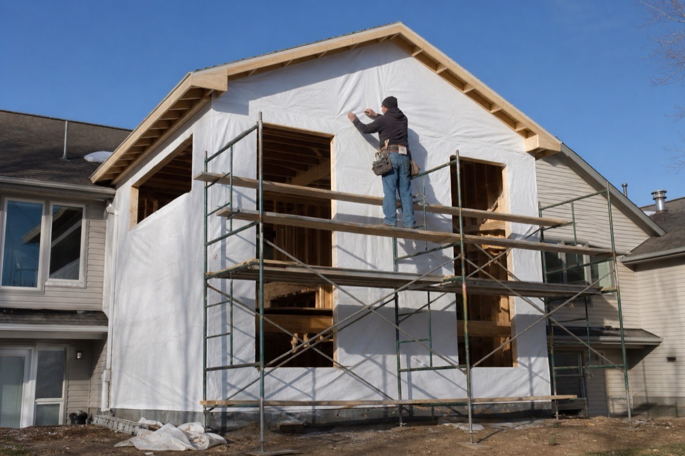 AI-generated illustration: a worker on scaffolding wraps a new two-story addition in plain house wrap.