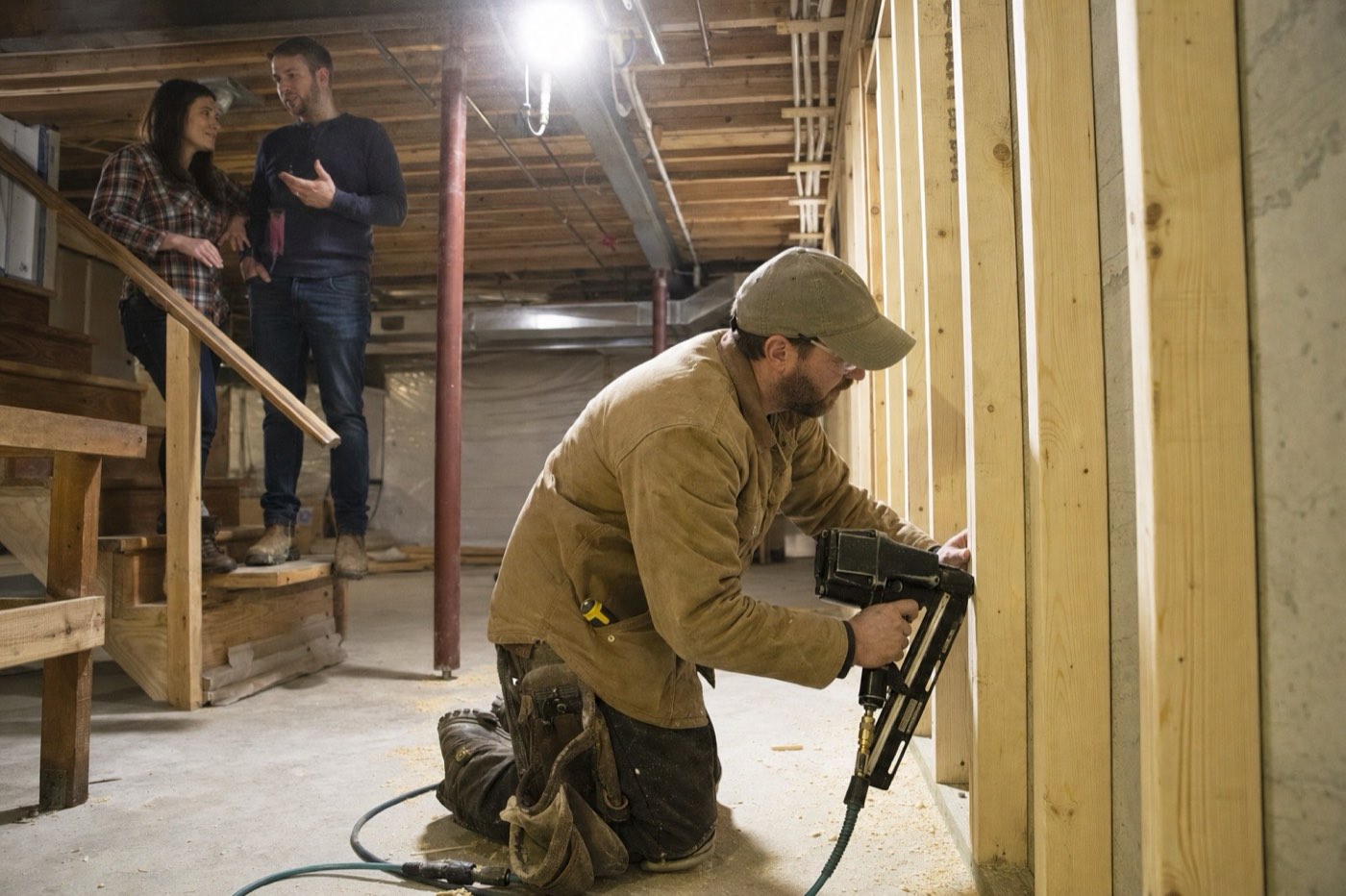 AI-generated illustration: a carpenter frames a basement wall with a nail gun while a couple talks on the basement stairs.