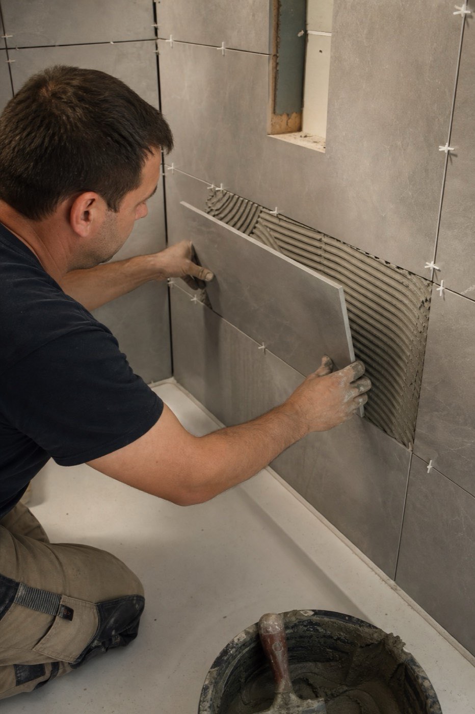 AI-generated illustration: a tile setter presses a large grey tile onto a shower wall.